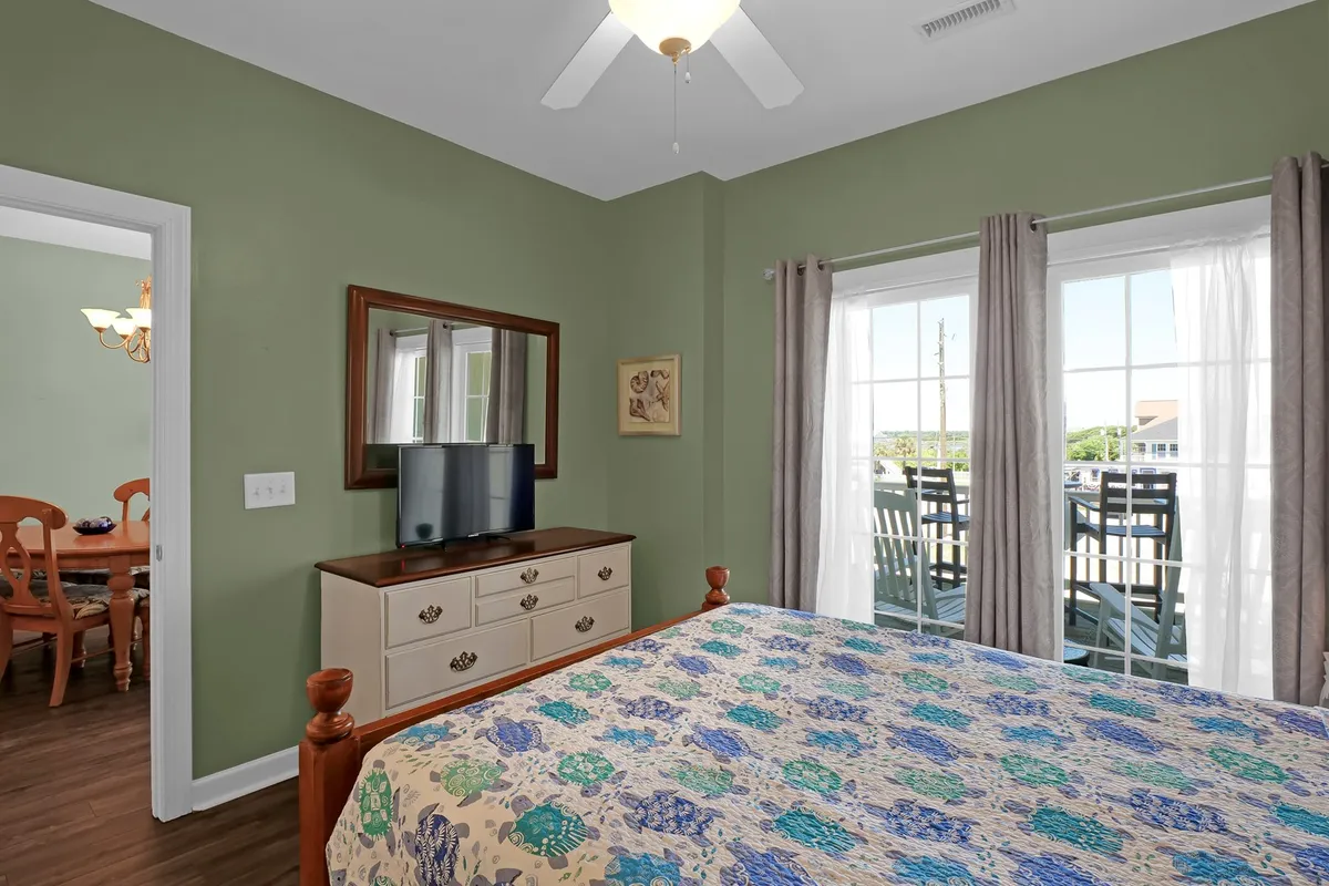 A cozy bedroom with colorful bedding, a dresser with a mirror, and large windows with curtains. The soft lighting and inviting atmosphere suggest personal comfort and the option to bring or rent linens, as well as a restful stay.