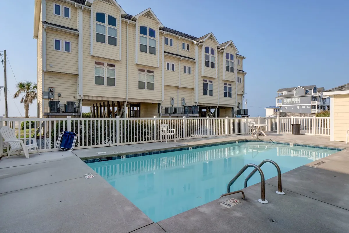 A resort-style swimming pool surrounded by palm trees and lounge chairs, with the beach and ocean just beyond. The pool area is inviting, offering both sunbathing spots and shady retreats, ideal for socializing or relaxing after a swim.