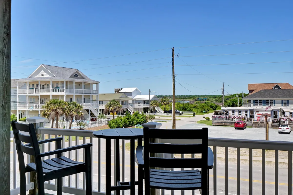 The photo features inviting balcony seating shaded by palm trees, overlooking buildings across the street—likely including nightlife venues. The scene suggests a relaxed vantage point for guests to enjoy sunset drinks, people-watch, and take in the neighborhood’s activity from the comfort of their condo balcony.