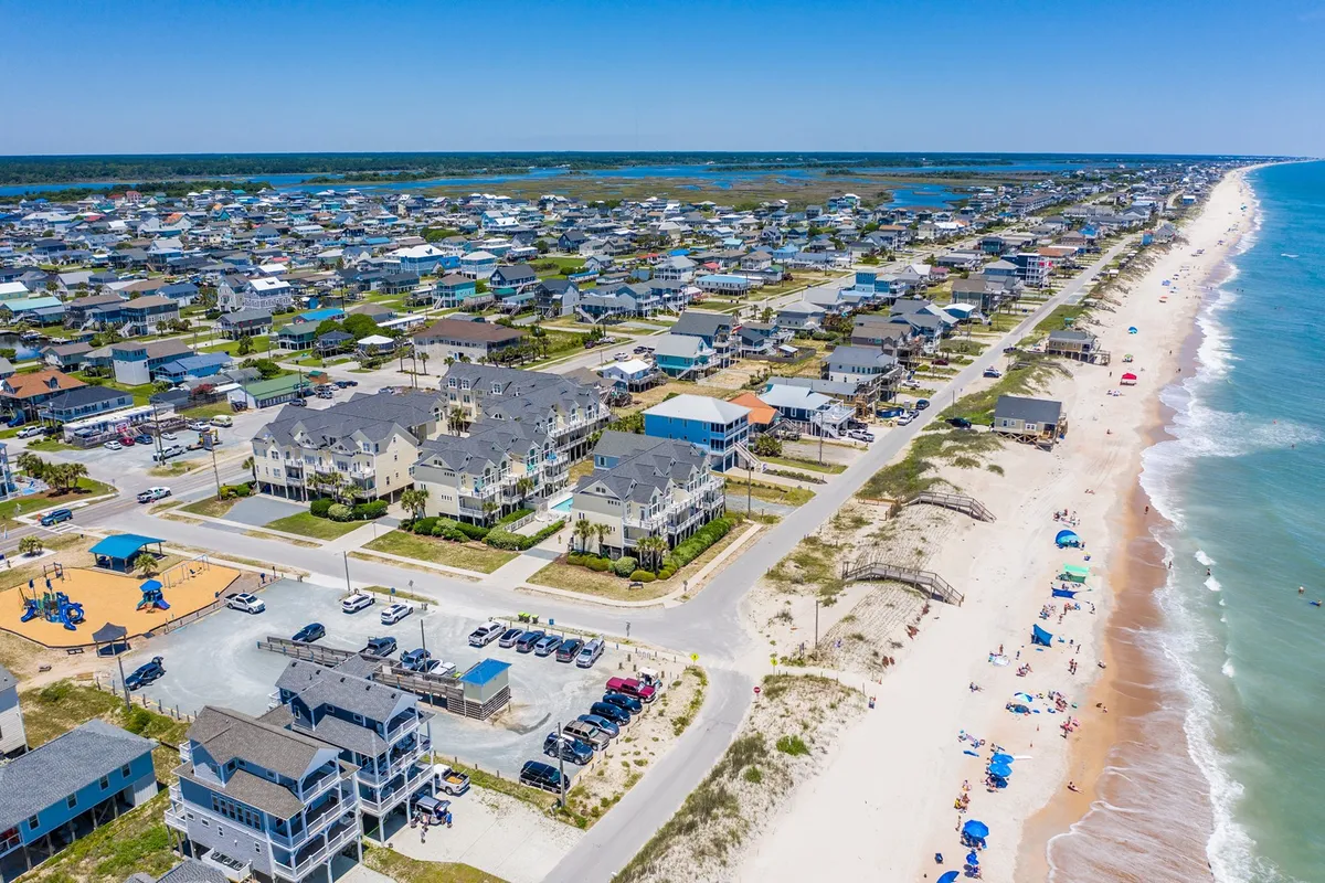 A wide-angle exterior shot showing a sandy beach, gentle ocean waves, seaside houses, a playground, and a few vehicles in the parking area. Families and children are playing, sunbathing, and enjoying a vibrant day by the shore with sandcastle-building and volleyball.