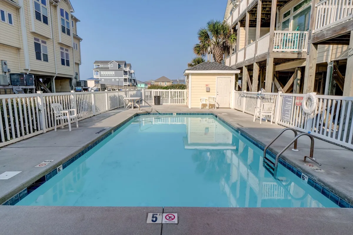 The resort-style outdoor swimming pool surrounded by lounge chairs and nearby buildings, with a lifeguard station visible. The scene is bright and relaxed, inviting guests to envision afternoons of affordable leisure and socializing just steps from their rental.
