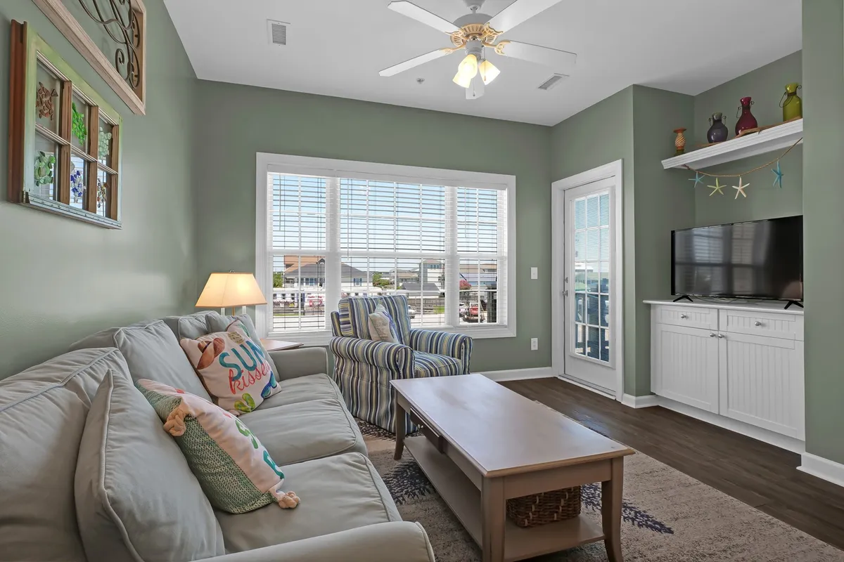 This cozy living room features a plush sofa with colorful cushions, striped armchairs, and a TV stand with decorative accents. A large window allows natural light to brighten the area, making it a perfect spot for relaxing or catching up after a day of beach adventures.