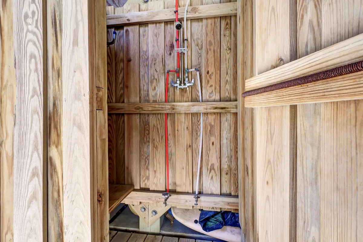 A rustic outdoor shower area with wooden walls, a wooden bench, and visible shower fixtures. The earthy tones and open-air feel evoke a refreshing experience, ideal for rinsing off after a sandy beach day.